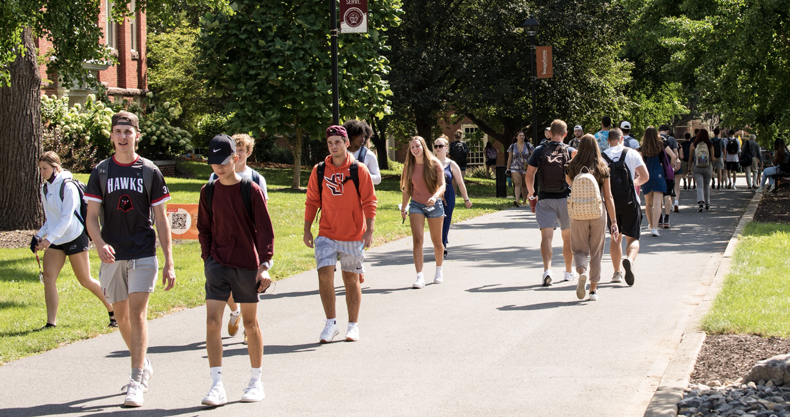 Groups of students walk along a tree-lined campus pathway on a sunny day, with many carrying backpacks and wearing casual summer clothing.